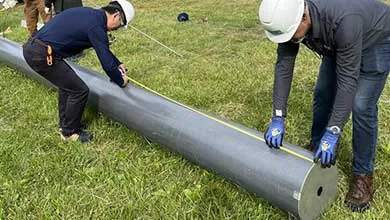 Two men in hard hats measure a large composite utility pole in a grassy field with a yellow tape measure.