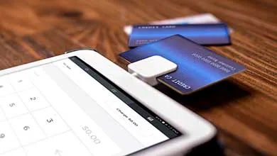 Blue credit card in a white square card reader attached to a tablet on a wooden surface.