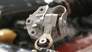 Gloved hands hold a metal car engine mount, engine bay blurred in the background.