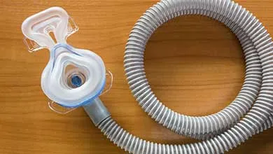 CPAP mask with clear and white cushion attached to a coiled, gray, corrugated hose on a wooden surface.