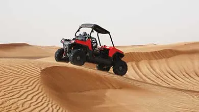 A red and black off-road buggy drives over a sand dune in a wide desert.