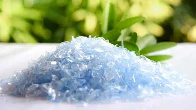 Close-up of a pile of light blue, translucent plastic flakes on a white surface, with blurred green leaves in the background.